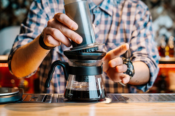 Fototapeta na wymiar Alternative Coffee Brewing Method. Close-up of the hands of barista who is preparing filter coffee in an aeropress. Barista wears a checkered flannel shirt and works at the bar counter.