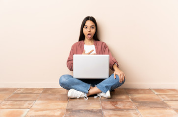 Young mixed race woman with a laptop sitting on the floor surprised and shocked while looking right