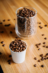 Two cups with roasted coffee beans - one transparent glass and one small paper cup on the black and brown wooden table in the restaurant