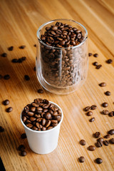 Two cups with roasted coffee beans - one transparent glass and one small paper cup on the black and brown wooden table in the restaurant
