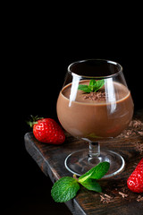 Chocolate pudding topped with mint in the glass against the black background. Ripe strawberry on the wooden board