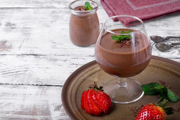 Chocolate pudding topped with mint in the glass. Ripe strawberry on the ceramic plate on the white wooden table