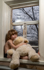 little boy with blond hair sitting on a windowsill holding on to a big soft toy