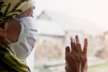 Fototapeta na wymiar An old grandmother in a medical mask looks through the window into the street. An elderly woman in a mask on her face on self-isolation during quarantine due to a coronavirus pandemic