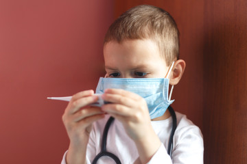 A young child doctor with a medical mask holds a thermometer in his hands and looks at the temperature. Close-up studio shot on a bright pink background for articles childhood diseases and covid-19