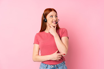 Young redhead woman over isolated pink background working with headset looking side