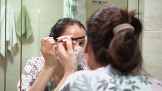 Pretty young brunette woman in protective medical pink mask in floral pajamas correcting shape of her eyebrows with a black pencil in front of the mirror at home. Life in quarantine of a coronavirus.
