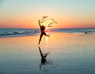 Young woman dancing with a ribbon on a beach at low tide, leaping into the air, silhouetted by a...
