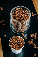 Two cups with roasted coffee beans - one transparent glass and one small paper cup on the black and brown wooden table in the cafe