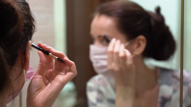 Beautiful brunette girl in floral blouse kimono in protective medical face mask correct shape of her eyebrows with a black in bathroom at home. Close focus on the hand. Covid19 virus isolation period.