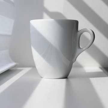White espresso coffee mug. Light effect, white and dark stripes fall on the subject and background. Light and shadow in the composition. A cup of coffee near the window.