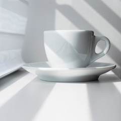 White espresso coffee mug. Light effect, white and dark stripes fall on the subject and background. Light and shadow in the composition. A cup of coffee near the window.