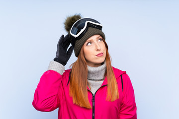 Skier redhead woman with snowboarding glasses over isolated blue wall having doubts and with confuse face expression