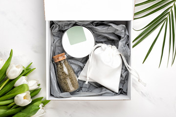 Cosmetic gift set of body or face cream container and bag with soap and a bottle of bath salts. Tulip flowers and palm leaf near box on white marble background, flat lay or top view - Powered by Adobe
