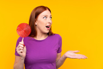 Young redhead woman holding a lollipop over yellow background with surprise facial expression