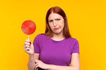 Young redhead woman holding a lollipop over yellow background with sad expression