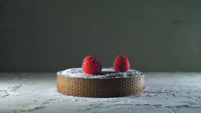 Chef serving a dark chocolate tart with sugar powder putting a raspberries on it, delicious dessert on grey background, close up