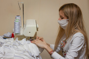 blonde girl with a white mask on her face sews gauze protective masks on an overlock sewing machine to protect against coronavirus during an epidemic