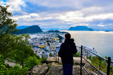 Fototapeta na wymiar Tourist man looks on the Alesund cityscape from Aksla viewpoint. Neoclassical and neo-Gothic stone buildings. Art Nouveau architecture. Sun rays illuminate Atlantic Ocean and islands. Norway.