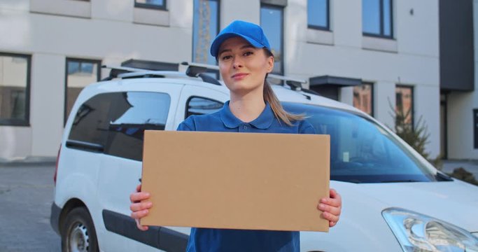 Portrait of young Caucasian beautiful woman in blue uniform and hat standing outdoor at white car with parcel in hands. Pretty female courier handing parcel to camera and smiling.