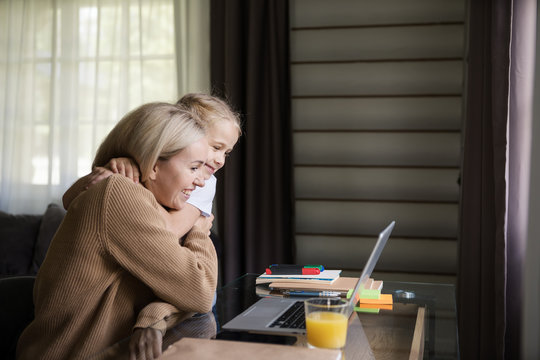 Side View Of Cute Little Daughter Hugging Her Happy Mother Sitting At Desk And Working On Laptop Computer From Home