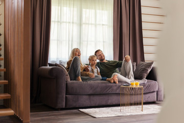 Excited family of mother, father and little daughter watching tv show together sitting on sofa at home