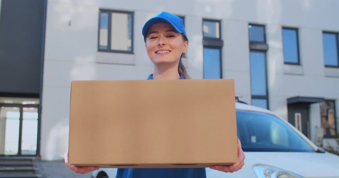 Portrait shot of young Caucasian cheerful woman in blue uniform and hat standing outdoors at white car with parcel in hands and smiling . Pretty female delivery worker handing parcel to camera.