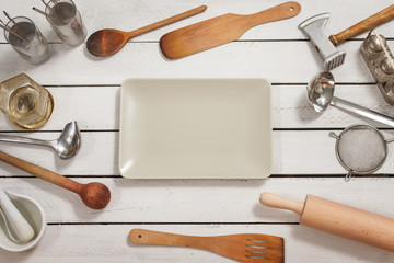 Prostokątna szklana biała miska leży na drewnianym stole. Na około porozkładane są stylowe naczynia kuchenne tworzące tło. - obrazy, fototapety, plakaty