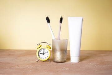 Toothbrushes in a glass and an alarm clock on a yellow background.