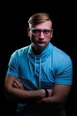Young man in glasses and a blue t-shirt stands with a pensive look on a black background.