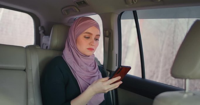 oung beautiful muslim woman in hijab in car passenger rear seat typing on cell phone with serious expression. Internet bank. Businesswoman working at smartphone in back seat of crowdsourced taxi