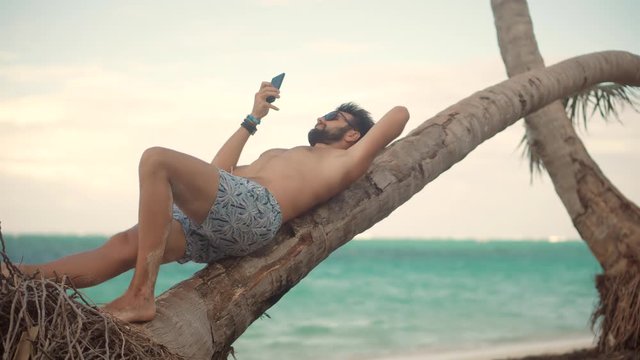 Attractive Tanned Man Sunbathing Using Mobile App For Photo Video.Selfie On Smartphone In Beautiful Place.Man Using Mobile Phone For Selfie Photo.Guy Taking Picture On Vacation Holiday Carribean Beach