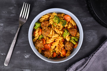 Bowl with meal made of fusilli pasta, broccoli and carrot vegetables and meatballs on dark table