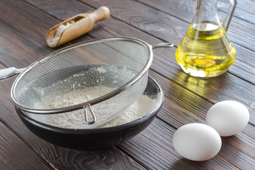 Sieve on black bowl with flour. Glass bottle with butter, eggs, wooden spoon with flour