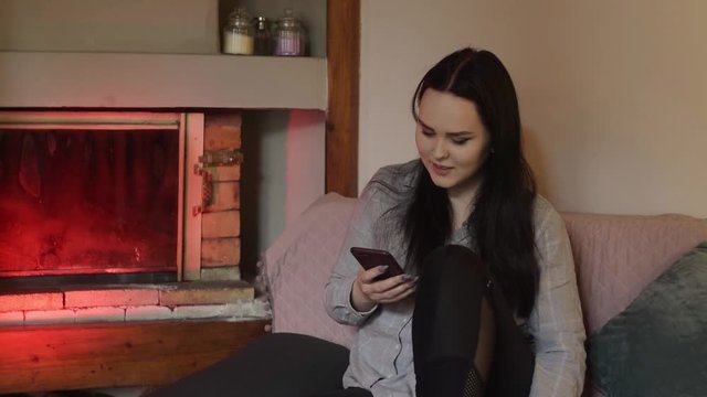 The brunette is sitting at home and looking at the phone, the girl is chatting, playing games, smiling. The girl sits dma near the fireplace, all the time on the phone.