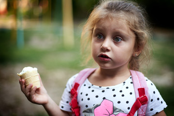 Portrait of a white-skinned girl with blond hair holding white ice cream in her hand. Mouth stained in ice cream. The girl is wearing a T-shirt and a pink backpack.