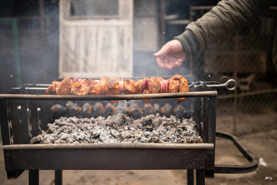A young man hand grilling a delicious shashlik (meat) and potatoes for a dinner in a metal outdoor braizer with grey ashes and smook