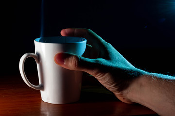 Taza de café blanca y caliente saliendo humo en la oficina de casa y mano que la sujeta para tomar...