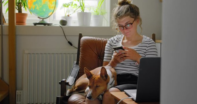 Young woman during break from work from home on laptop. Pretty woman sit in cosy comfortable armchair scroll through social media on smartphone. Bored and cosy at home with pet dog during lockdown