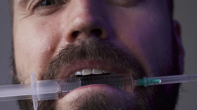 Macro: Angry man scornfully looks into the camera and holds a syringe in his teeth