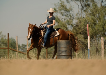 Cowgirl in cowboy hat riding brown horse through outside dirt arena, barrel racing pattern practice...