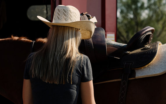 Western lifestyle image of cowgirl in straw cowboy hat close up, looking away while putting saddle on horse.