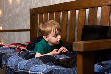 Fototapeta na wymiar a small child lies on a sofa in an apartment and carefully looks into a laptop. quarantined online training.