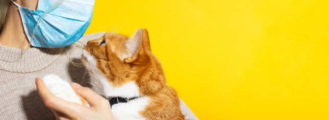 Close-up of young girl wearing medical mask against coronavirus, holding red white cat on yellow background with copy space.
