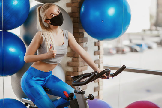 A pretty girl in a protective mask is engaged in an exercise bike. Protective masks against virus infection. Training during quarantine in the gym. Coronavirus (COVID-19) protection concept. - Powered by Adobe