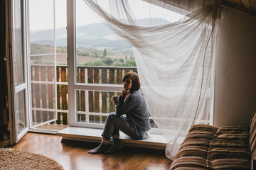 Young woman in woolen knitted sweater relaxing near window in cozy wooden cottage with mountains view.