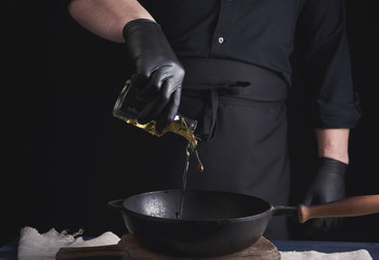 chef in black latex gloves pours olive oil from a transparent bottle into a black cast-iron frying...