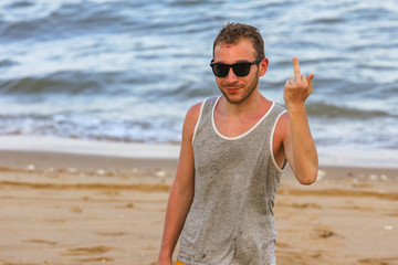 Fototapeta na wymiar young bearded man shows middle finger while looking at the camera (on background of ocean)