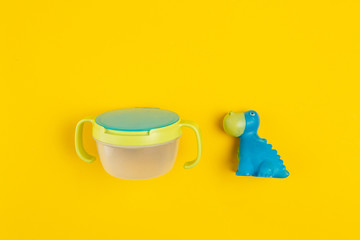 Obraz na płótnie Canvas Baby accessories. Infant Snacks cup and rubber dinosaur on yellow background.