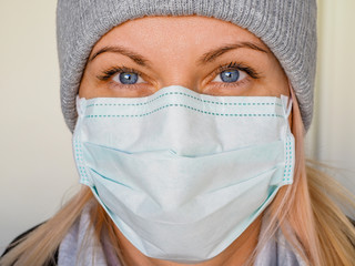 The face of a young beautiful blonde woman in a gray hat and a medical mask of blue or green color on a light backgroud. Health prevention during isolation due coronavirus epidemic. Healthy lifestyle.